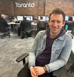Josh sitting in the office with 4 colleagues sitting at screens behind him