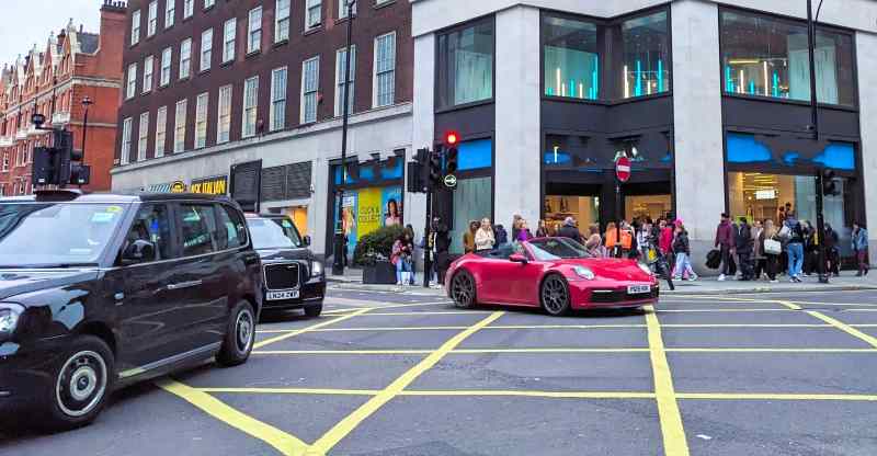 red porsche car entering box junction. Black cabs on left