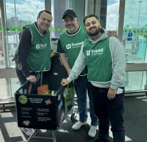 3 people wearing green bibs standing behind a supermarket tolley